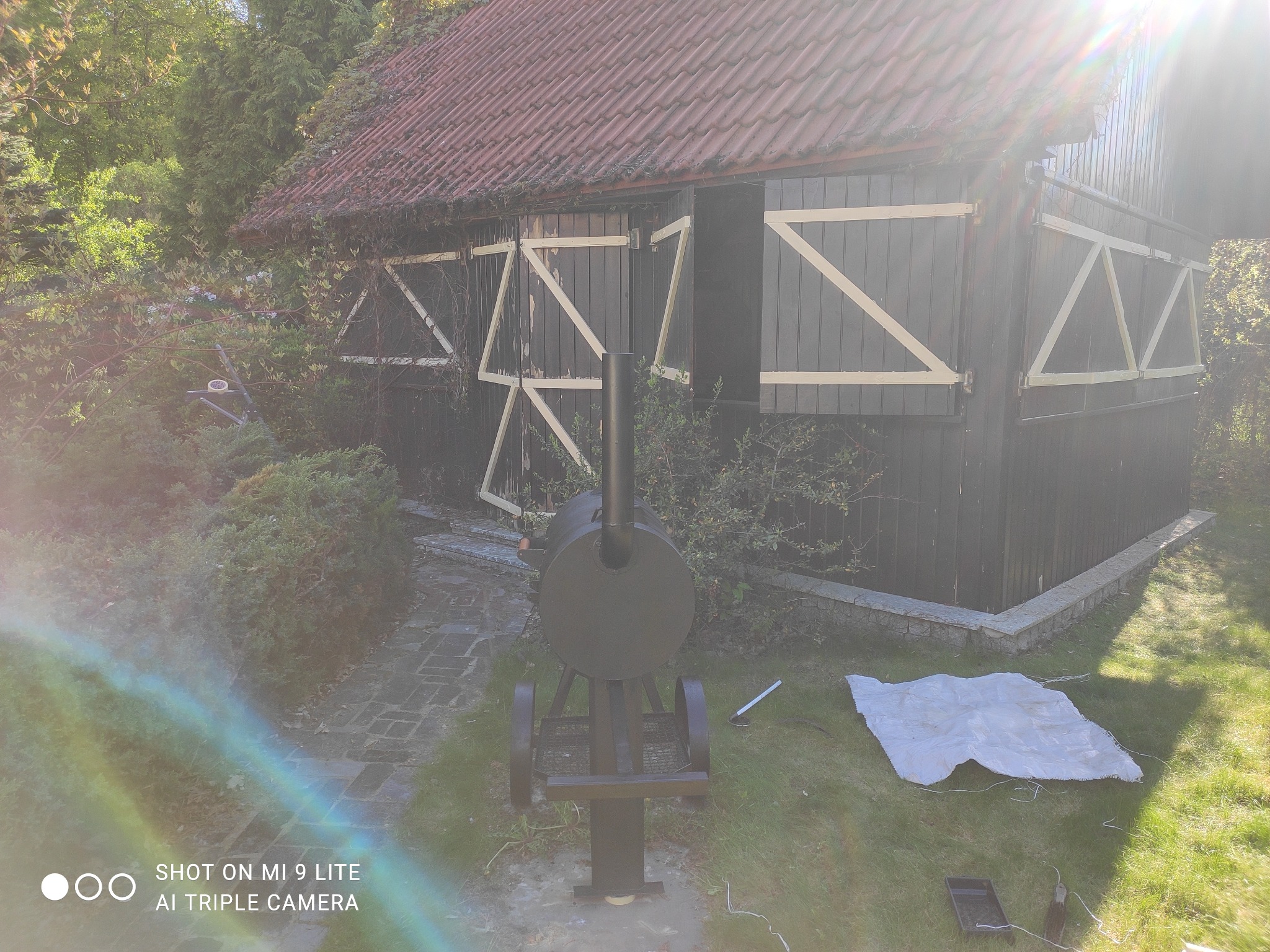 Rustykalna szopa z czerwonym dachem i czarnymi ścianami z dekoracyjnymi, jasnymi belkami, obok której stoi czarny grill na kółkach, a na trawie leży biała plandeka.