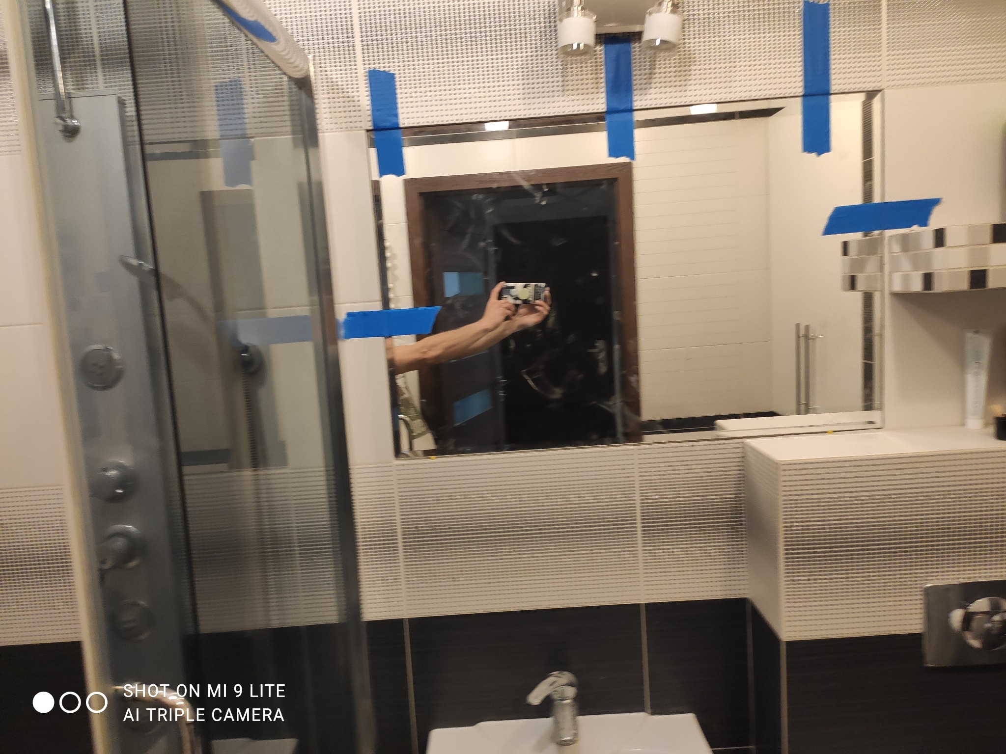 Łazienka w trakcie remontu z prysznicem, umywalką i lustrem zabezpieczonym niebieską taśmą malarską, widoczne odbicie osoby robiącej zdjęcie.