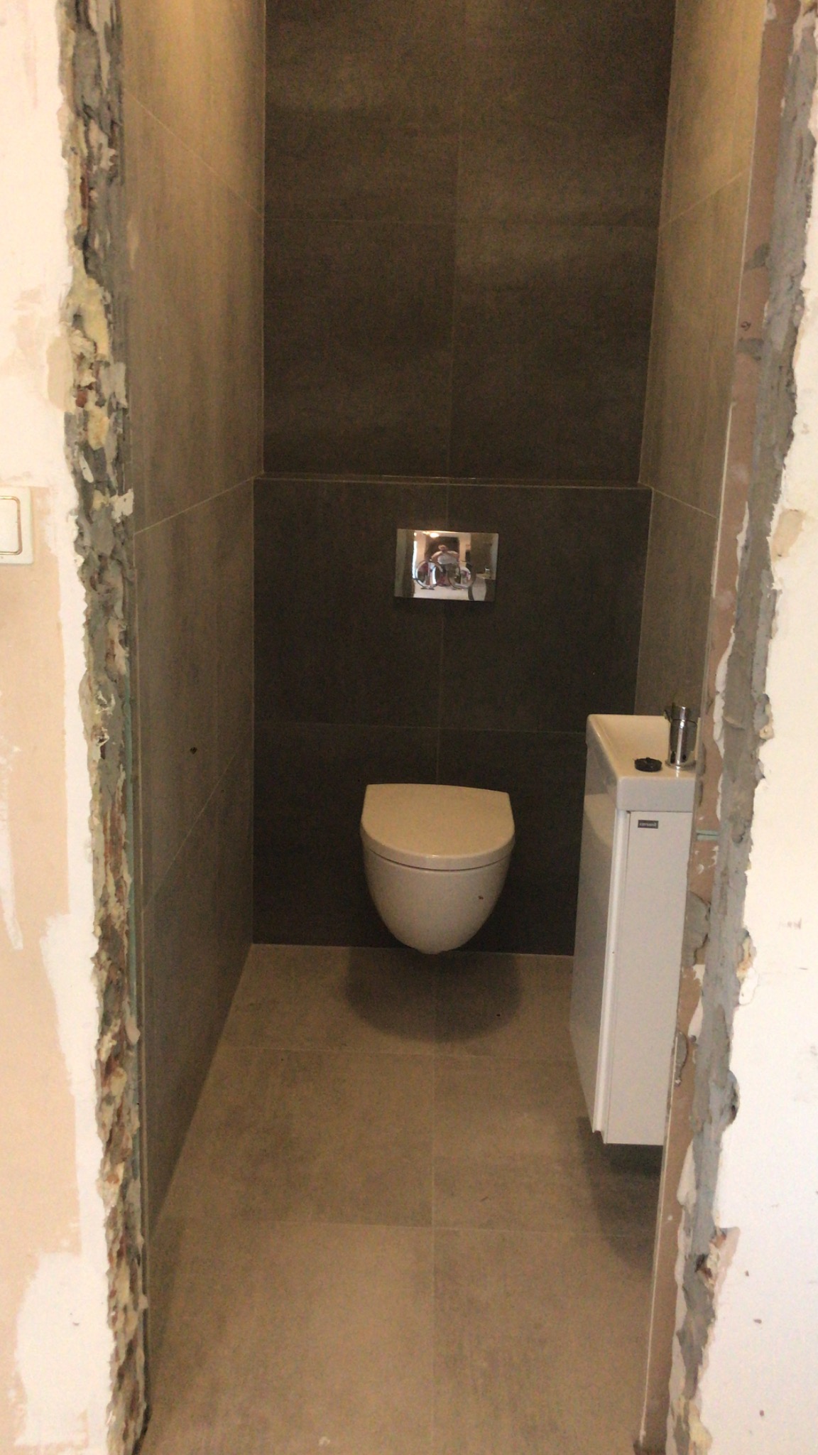 Wnętrze małej łazienki w trakcie wykańczania, z podwieszaną toaletą, umywalką narożną i ciemnymi płytkami na ścianach, widoczne surowe wykończenie ścian przy drzwiach.