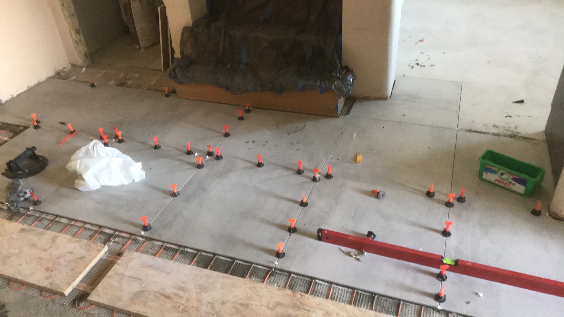 Szare płytki podłogowe w trakcie układania, z widocznym ogrzewaniem podłogowym, poziomnicą i systemem poziomowania płytek, w tle niezabezpieczony mebel pod folią.