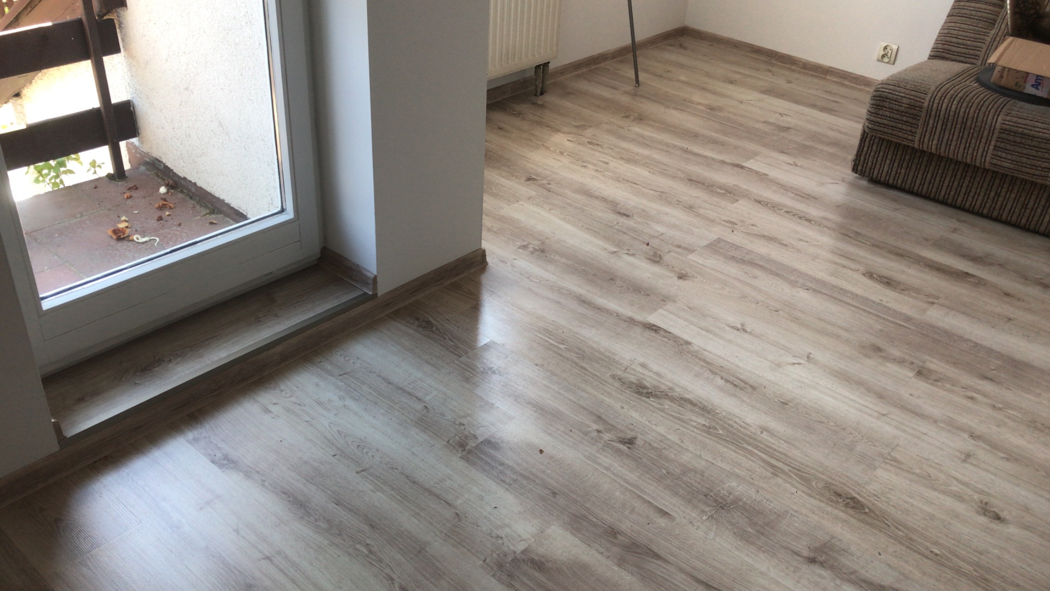 Ułożona podłoga z paneli laminowanych o jasnym odcieniu drewna, widoczny fragment balkonu z białymi drzwiami balkonowymi i fragment sofy w tle.