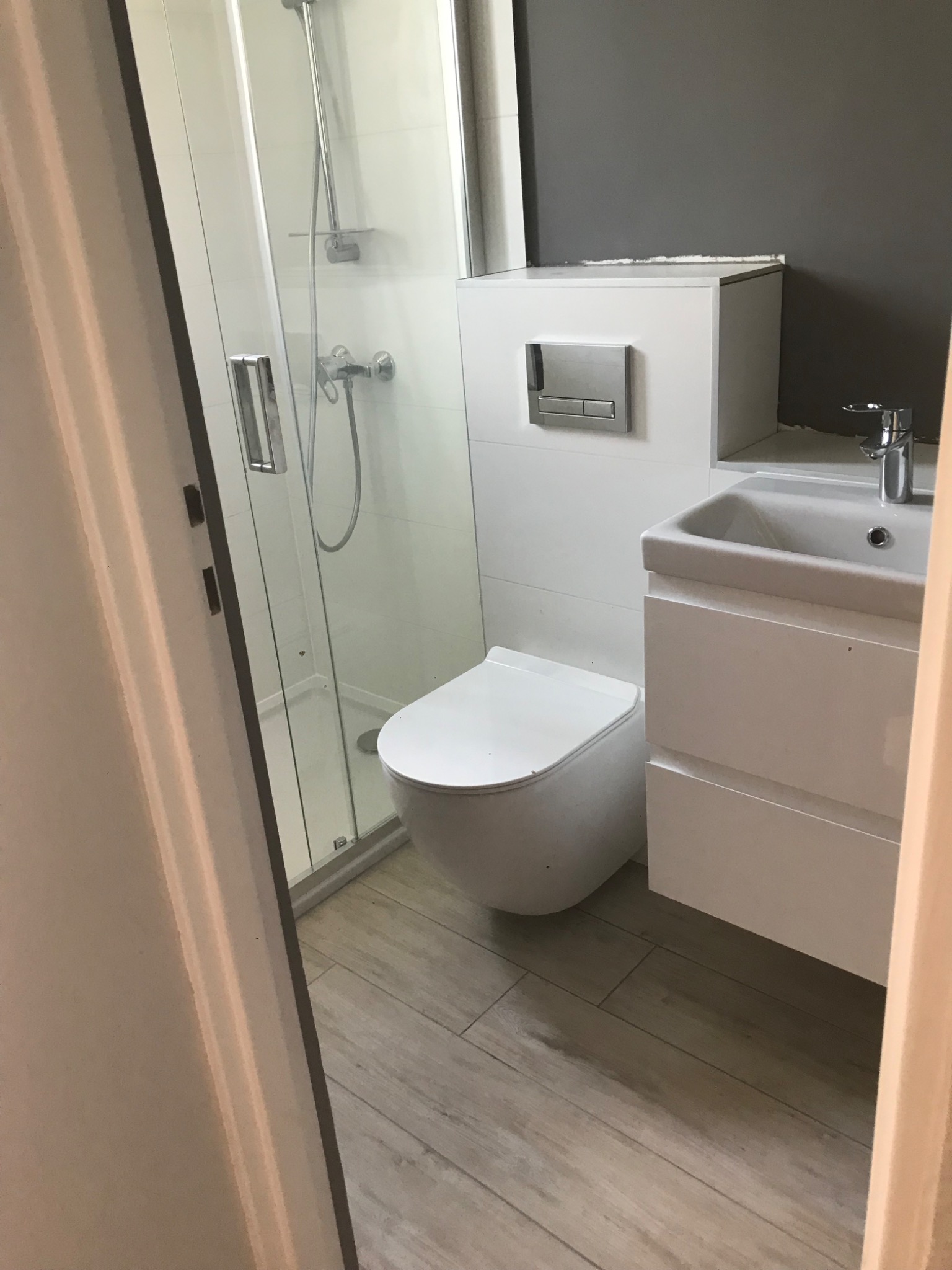 Nowoczesna łazienka z kabiną prysznicową ze szklanymi drzwiami, podwieszaną toaletą i białą szafką z umywalką na drewnopodobnych płytkach podłogowych, widziana zza framugi drzwi.