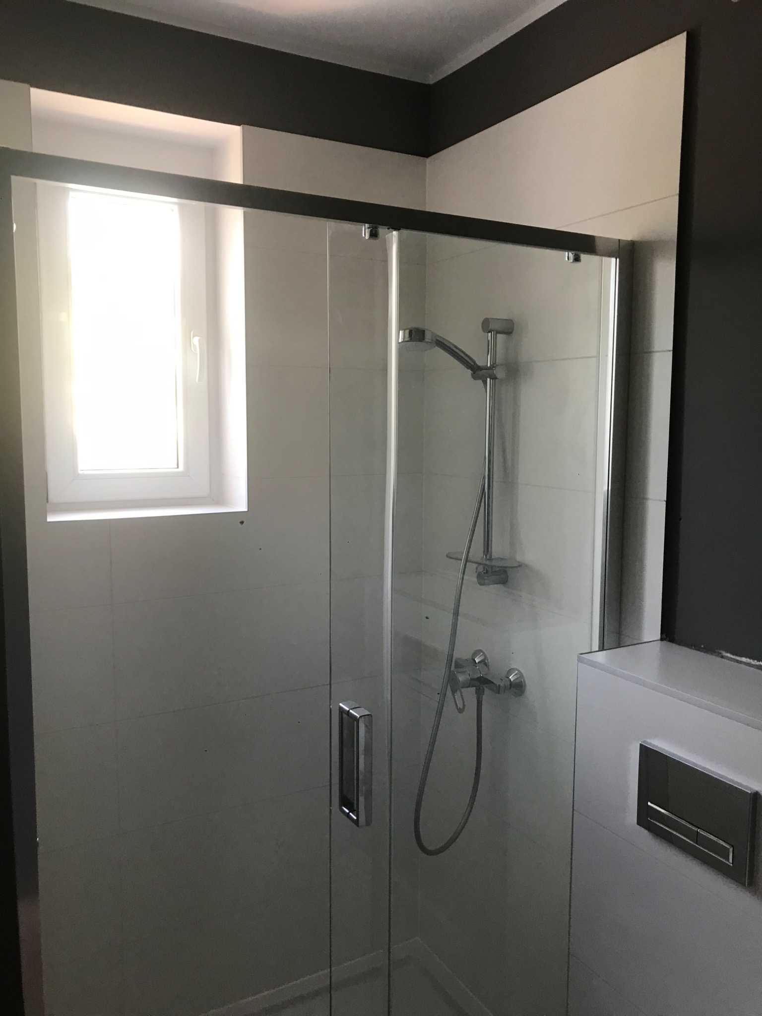 Nowoczesna kabina prysznicowa z ciemną ramą, widoczna armatura, białe płytki i okno w tle, z ciemnoszarym akcentem na suficie.