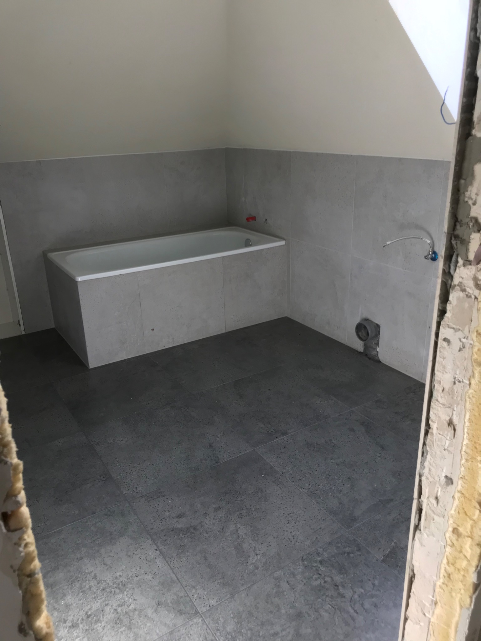 Wnętrze łazienki w trakcie remontu, z zamontowaną wanną obudowaną szarymi płytami, szara glazura na ścianach, widoczny fragment obitej futryny drzwi i szara podłoga z płytek.