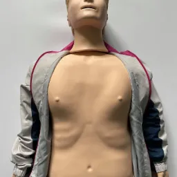 Manekin treningowy do nauki resuscytacji krążeniowo-oddechowej, ubrany w rozpiętą kurtkę sportową z różowym zamkiem.