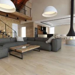 Przestronny salon z szarą sofą narożną, marmurowym stolikiem kawowym i wiszącym kominkiem, oświetlony dużymi, okrągłymi lampami. W tle widoczna kuchnia i schody z kamienną ścianą.