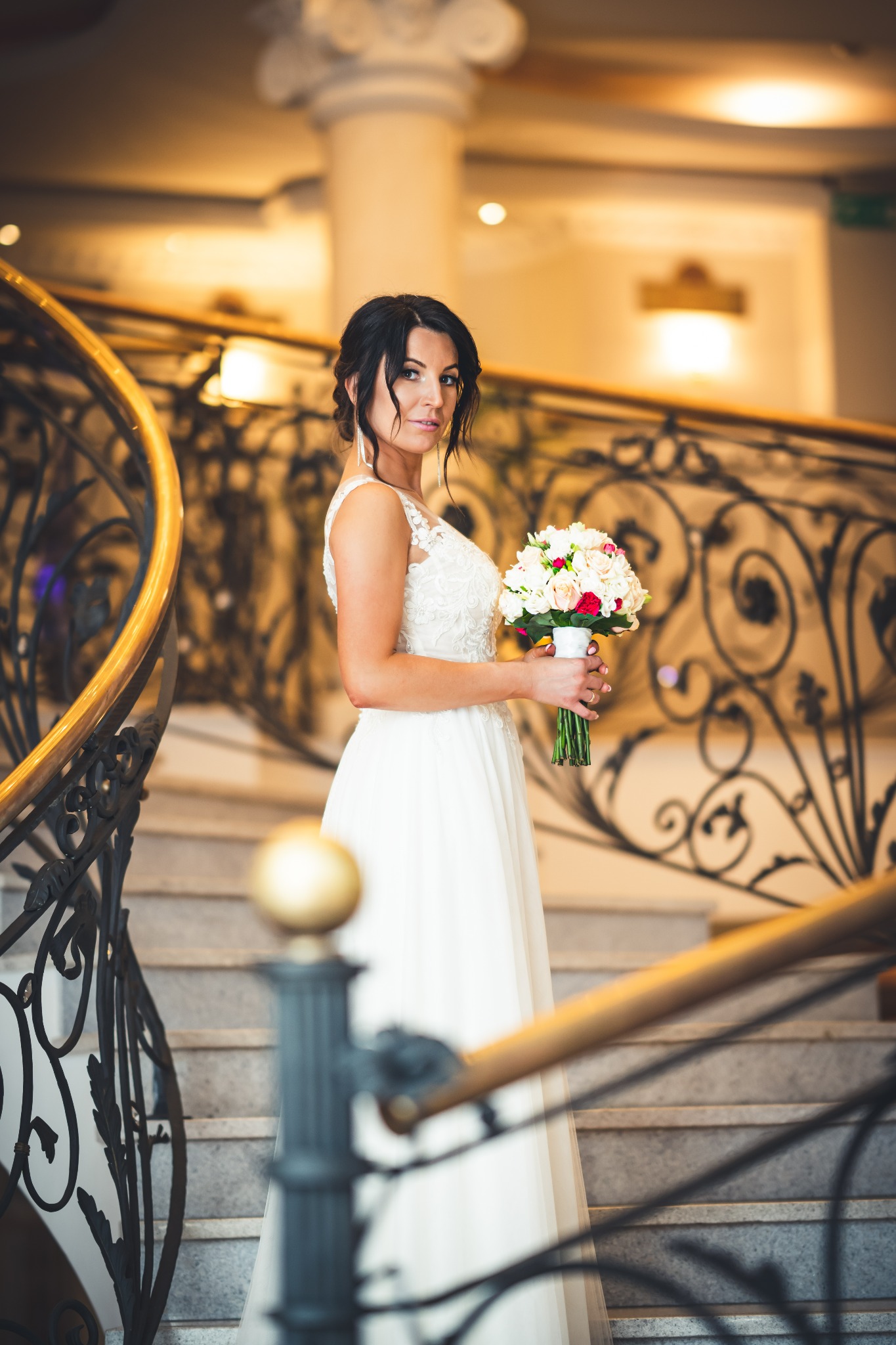 Panna młoda w białej sukni z bukietem stoi na marmurowych schodach z kutą, zdobioną balustradą, spoglądając w obiektyw.