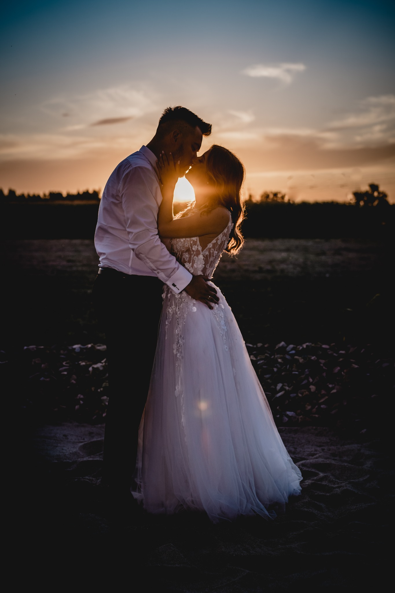 Silhouettes całującej się pary młodej na tle zachodzącego słońca, ona w białej sukni z koronkowymi aplikacjami i odkrytymi plecami, on w białej koszuli z podwiniętymi rękawami i ciemnych spodniach.