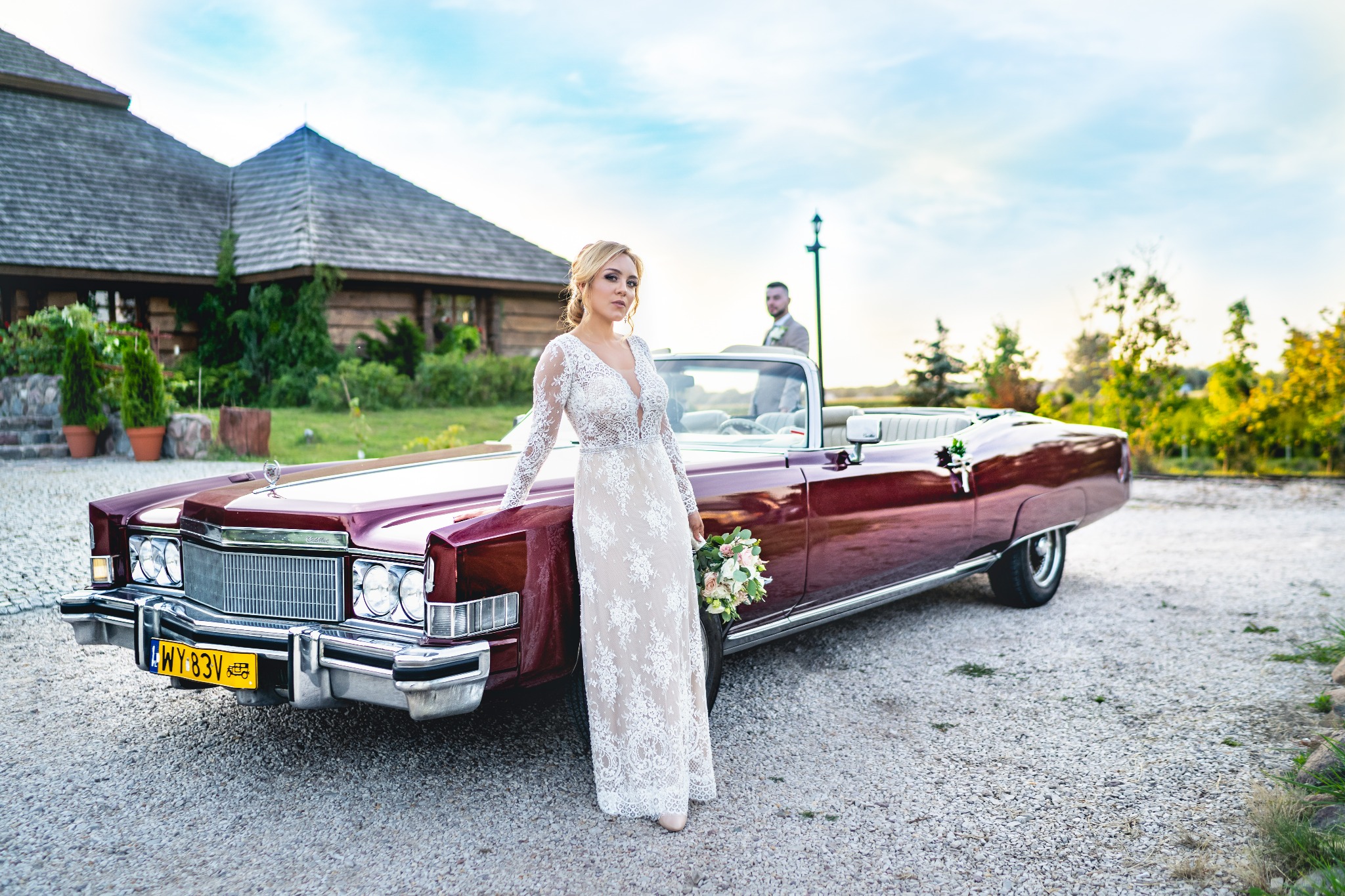 Panna młoda w koronkowej sukni opiera się o bordowego, zabytkowego kabrioleta; w tle drewniany budynek i pan młody.