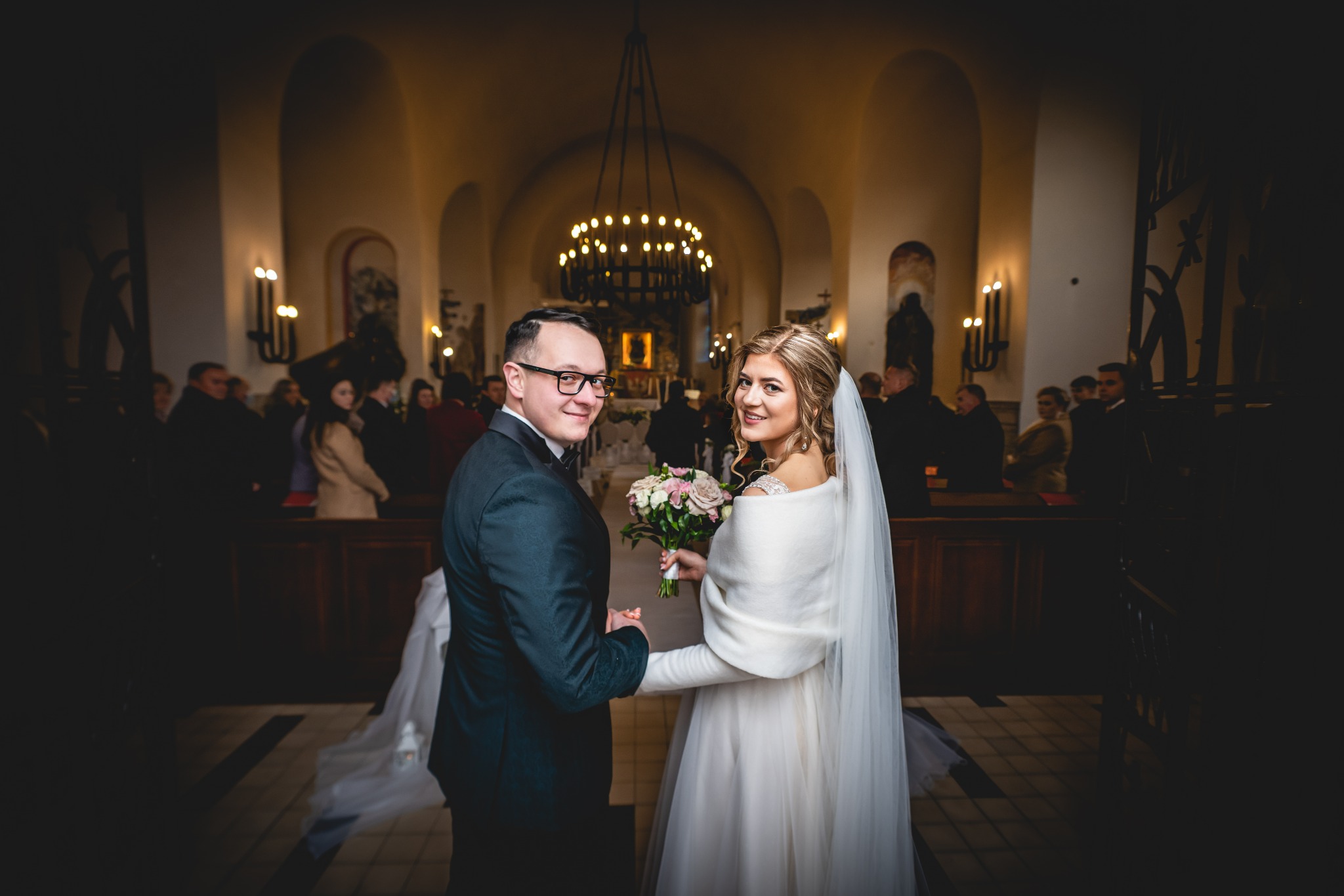 Para młoda trzymająca się za ręce, w kościele z żyrandolem i obserwującymi gośćmi.