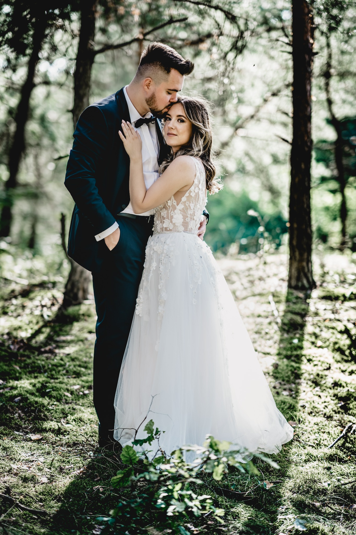 Para młoda w lesie: panna młoda w koronkowej sukni ślubnej z odkrytymi ramionami, pan młody w granatowym garniturze i muszce, delikatne światło słoneczne przebija się przez drzewa.