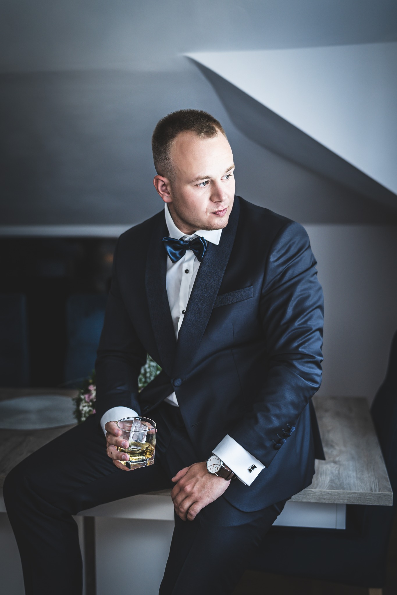 Mężczyzna w granatowym smokingu z muchą, siedzący na blacie, trzyma szklankę z whisky i lodem, na nadgarstku widoczny zegarek.
