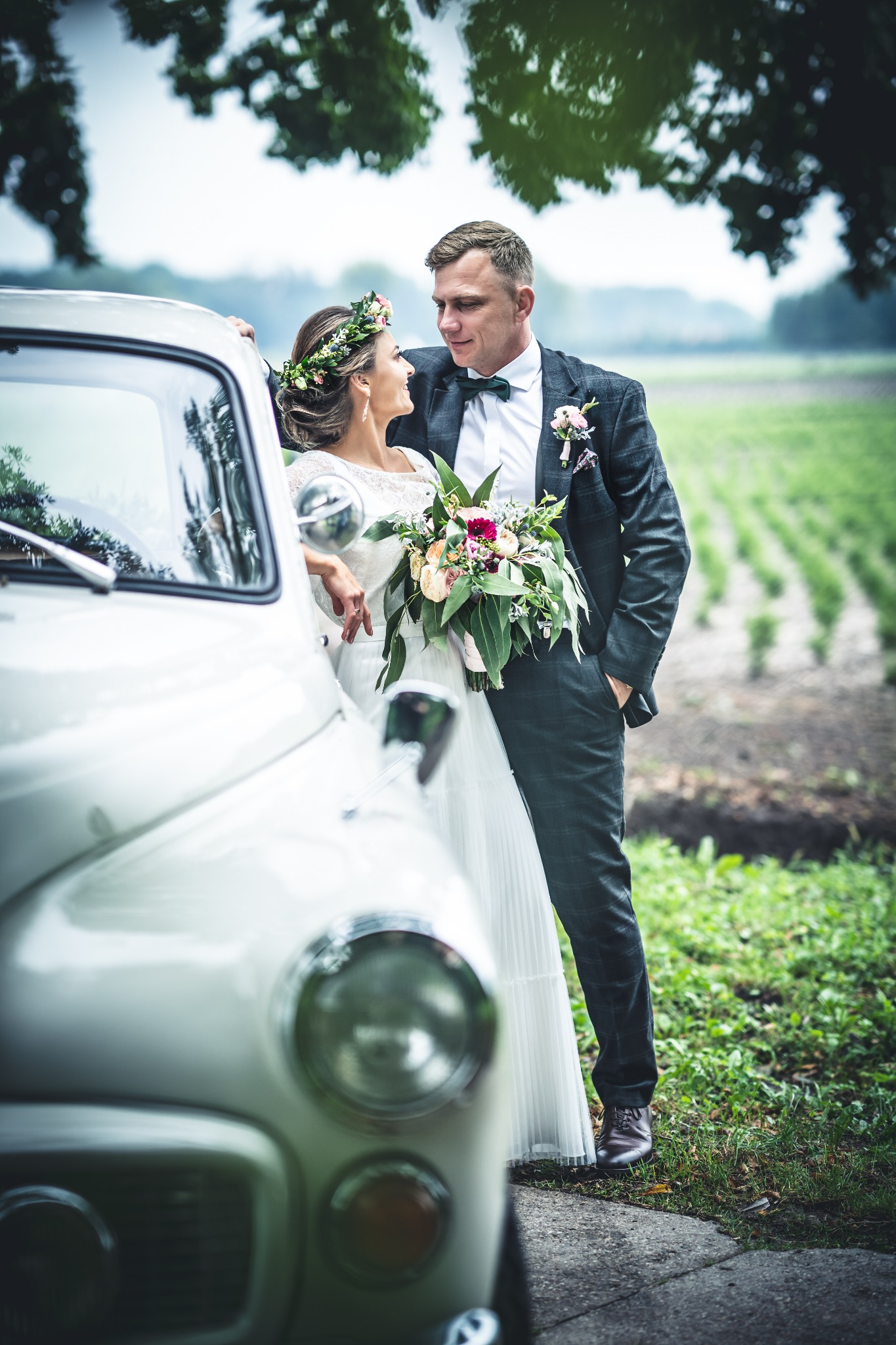 Para młoda wpatrzona w siebie, opierająca się o biały, zabytkowy samochód na tle zielonego pola.