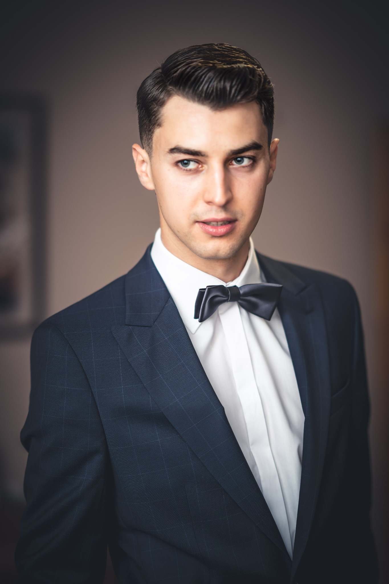 Portret eleganckiego mężczyzny w granatowym garniturze i czarnej muszce, z delikatnym uśmiechem i spojrzeniem w bok.