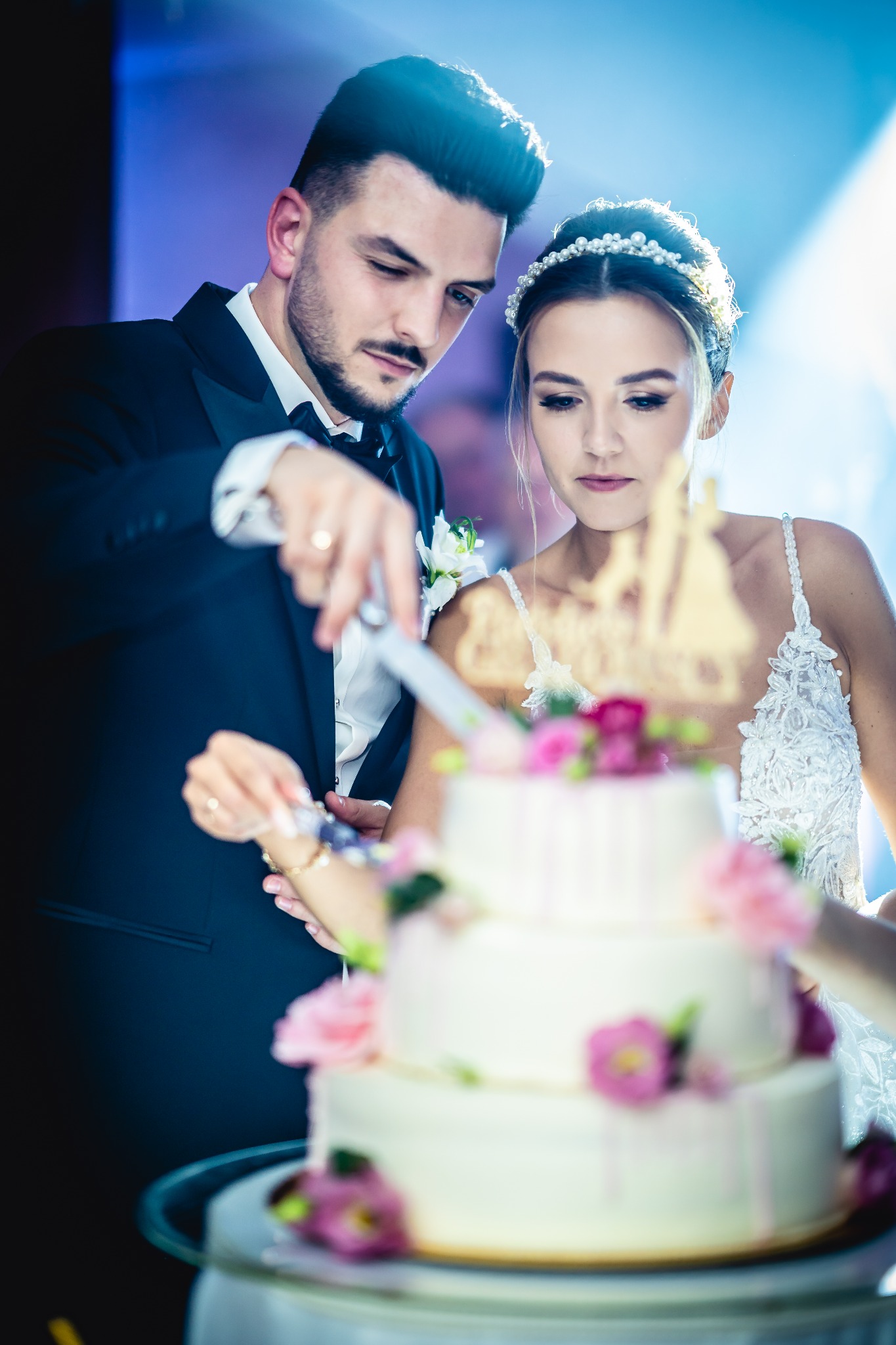 Młoda para kroi tort weselny ozdobiony różowymi kwiatami i figurką pary młodej, on w czarnym smokingu, ona w białej sukni z koronką i perłowym diademem.