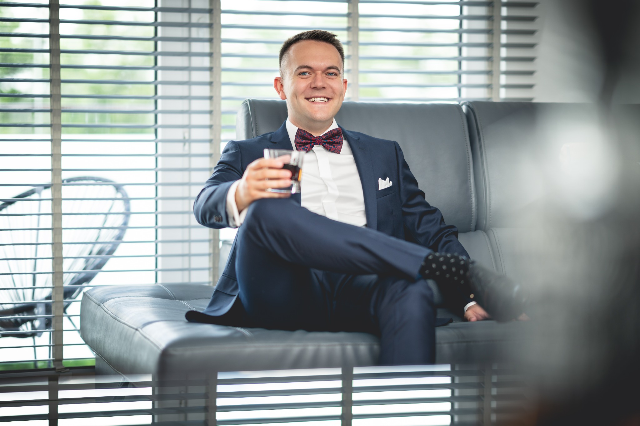 Uśmiechnięty mężczyzna w granatowym garniturze i bordowej muszce siedzi na szarej skórzanej sofie z drinkiem w ręku, w tle żaluzje okienne.