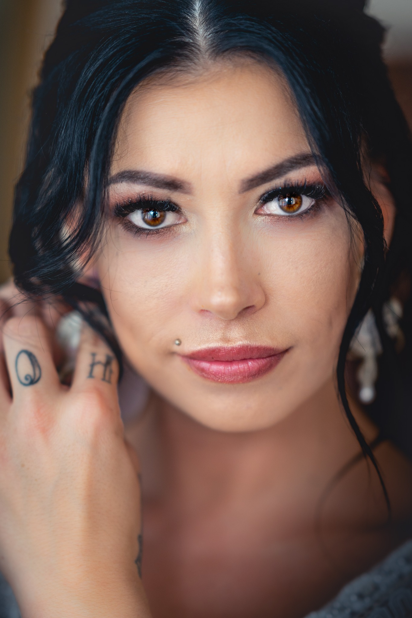 Portret kobiety z ciemnymi włosami, brązowymi oczami i tatuażami na dłoni, delikatny makijaż, kolczyk nad ustami.