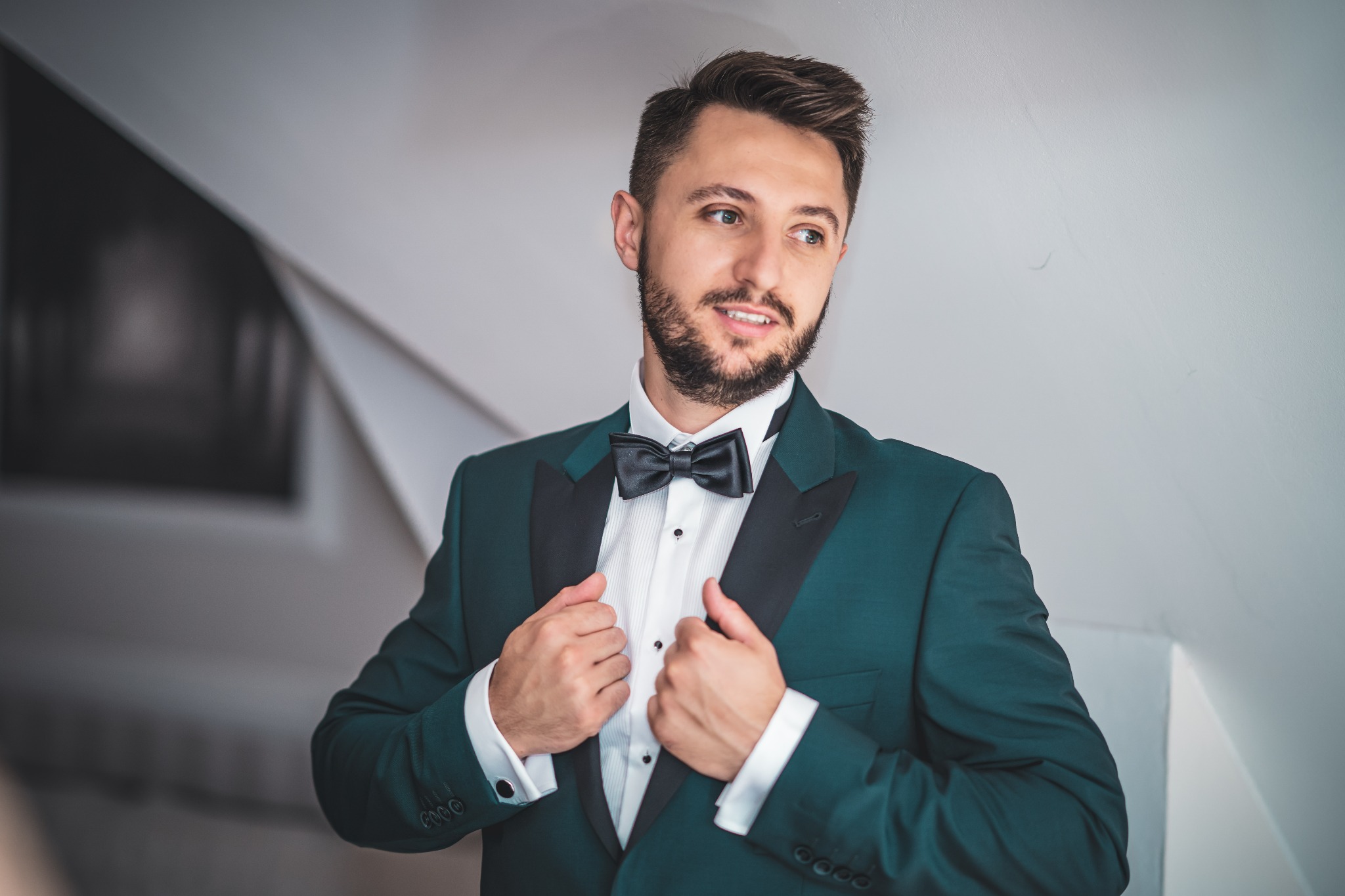 Elegancki mężczyzna w ciemnozielonym smokingu z czarną muchą, pozujący na tle jasnych ścian z subtelnym cieniem.
