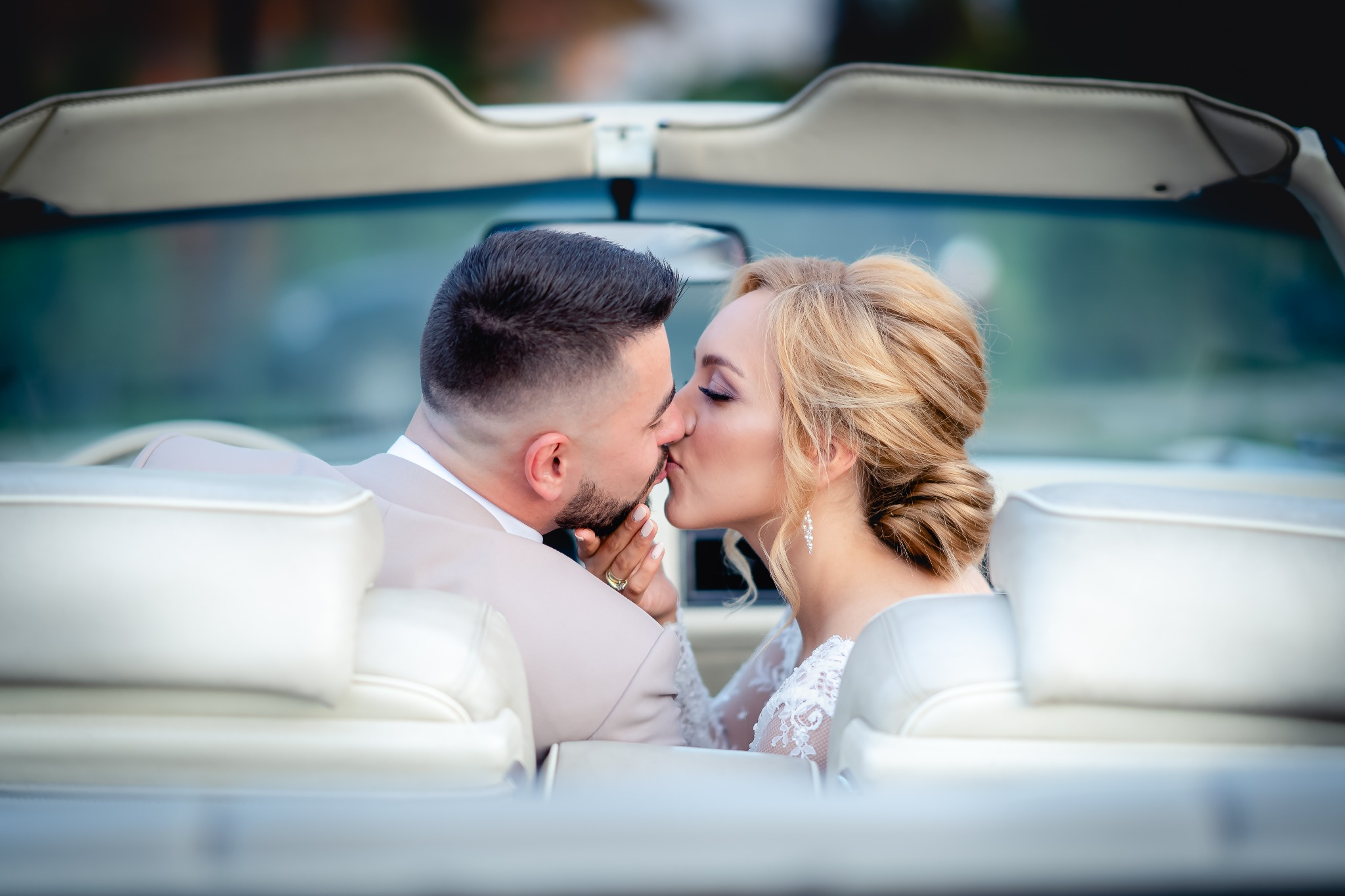 Pocałunek nowożeńców w białym kabriolecie z jasną skórzaną tapicerką, ona w koronkowej sukni, on w beżowym garniturze, widok zza siedzeń.