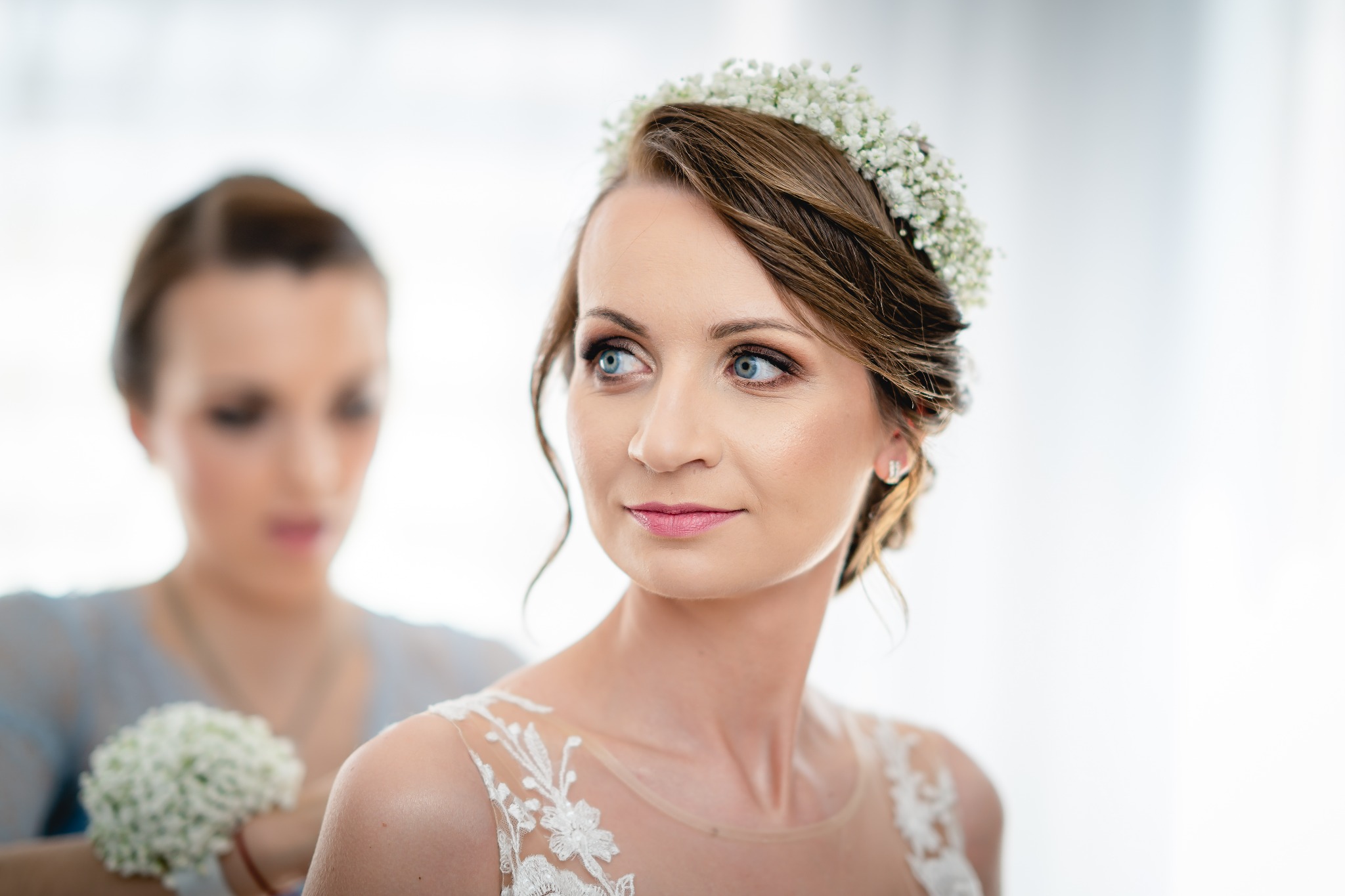 Panna młoda z wiankiem z gipsówki spogląda w bok podczas przygotowań, w tle asystentka z bukietem.