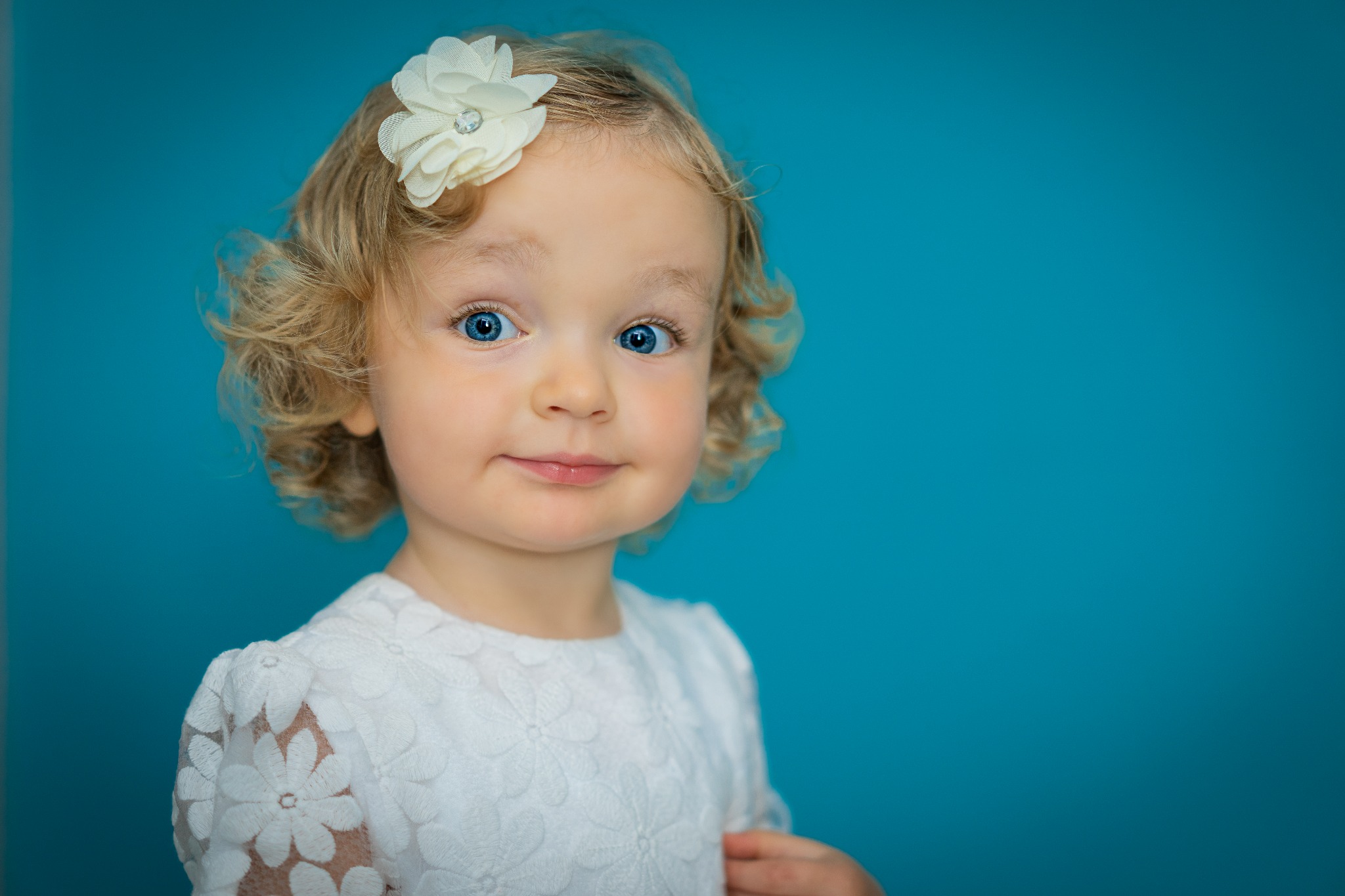 Portret uroczej dziewczynki z Sochaczewa o dużych niebieskich oczach i blond lokach, ozdobionych białą kwiatową spinką, na jednolitym turkusowym tle.