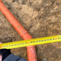 Geomatic Geodezja Mateusz Laskowski - Pomarańczowa rura kanalizacyjna w wykopie mierzona żółtą taśmą mierniczą, widoczny fragment ubrania osoby mierzącej i but w ziemi.