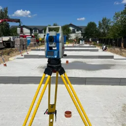 Żółty niwelator na trójnogu ustawiony na betonowej płycie fundamentowej w trakcie budowy domu jednorodzinnego, w tle pracujący robotnicy i dźwig podnoszący palety z cegłami.