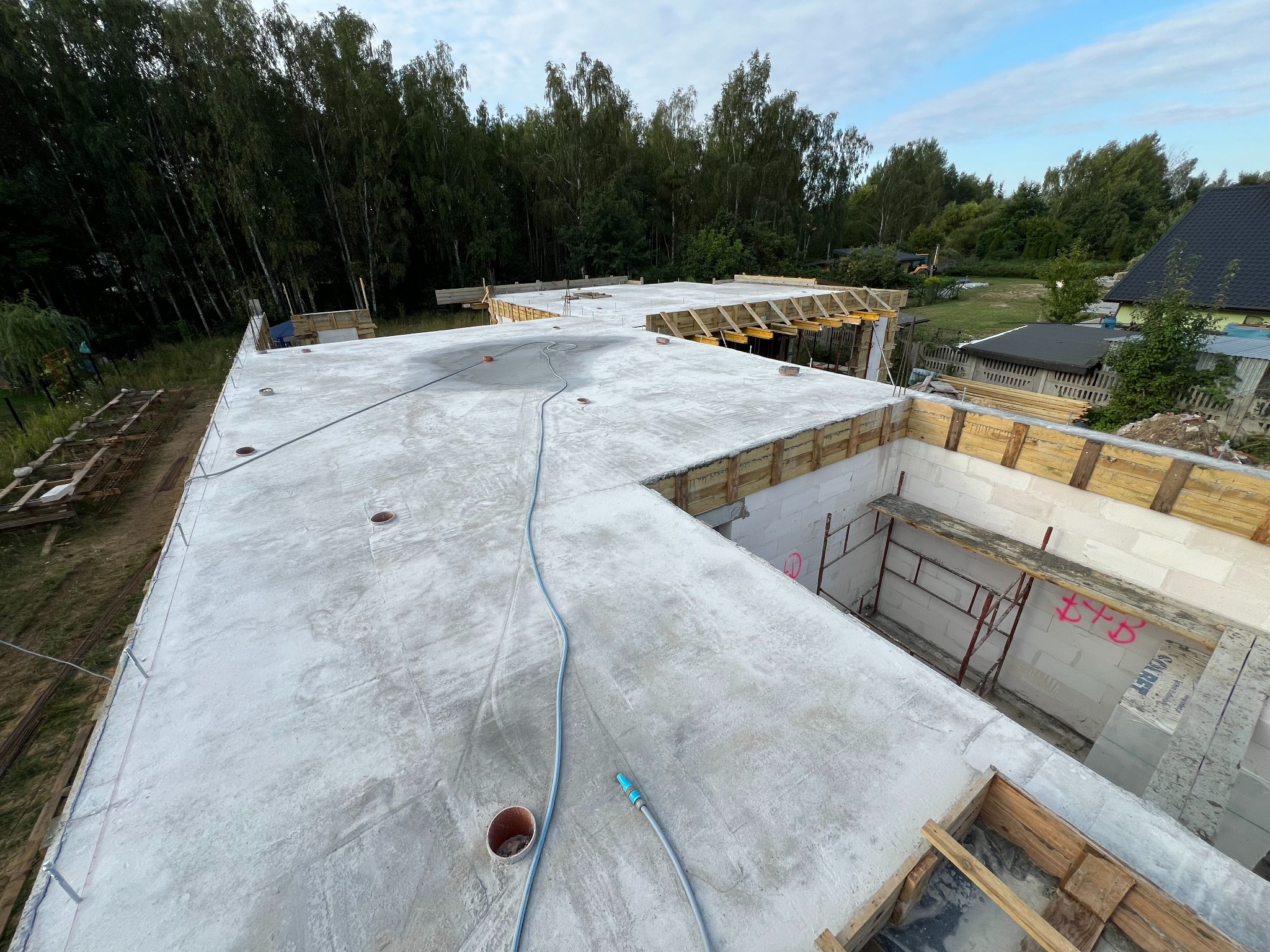 Wylany betonowy strop na budowie domu jednorodzinnego, widoczne szalunki i fragment ściany z białych bloczków, w tle drzewa.