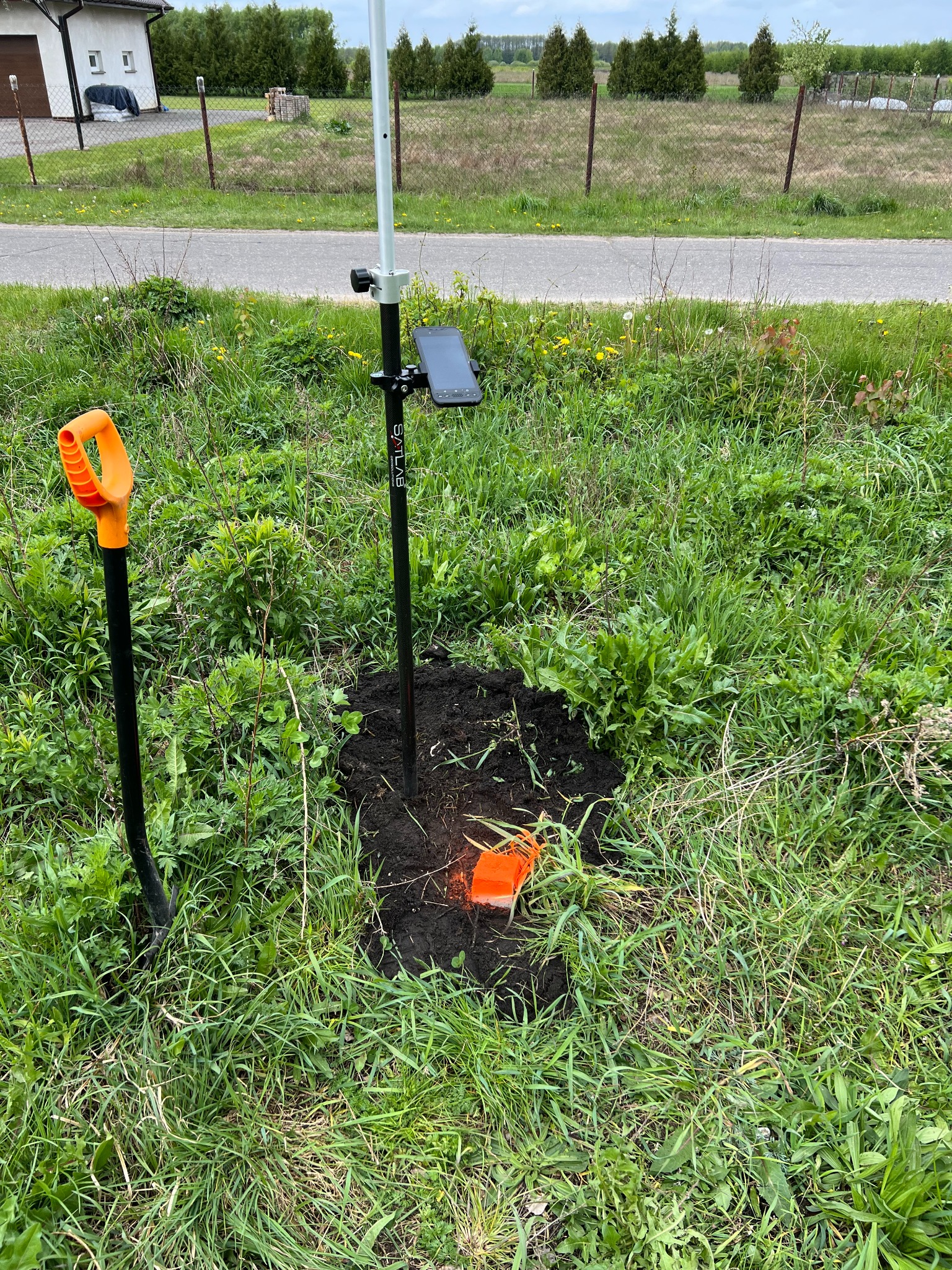 Pomiar geodezyjny w terenie: czarny statyw z tabletem, łopata z pomarańczową rączką i pomarańczowy znacznik w świeżo przekopanej ziemi na tle trawy, domu i ogrodzenia.