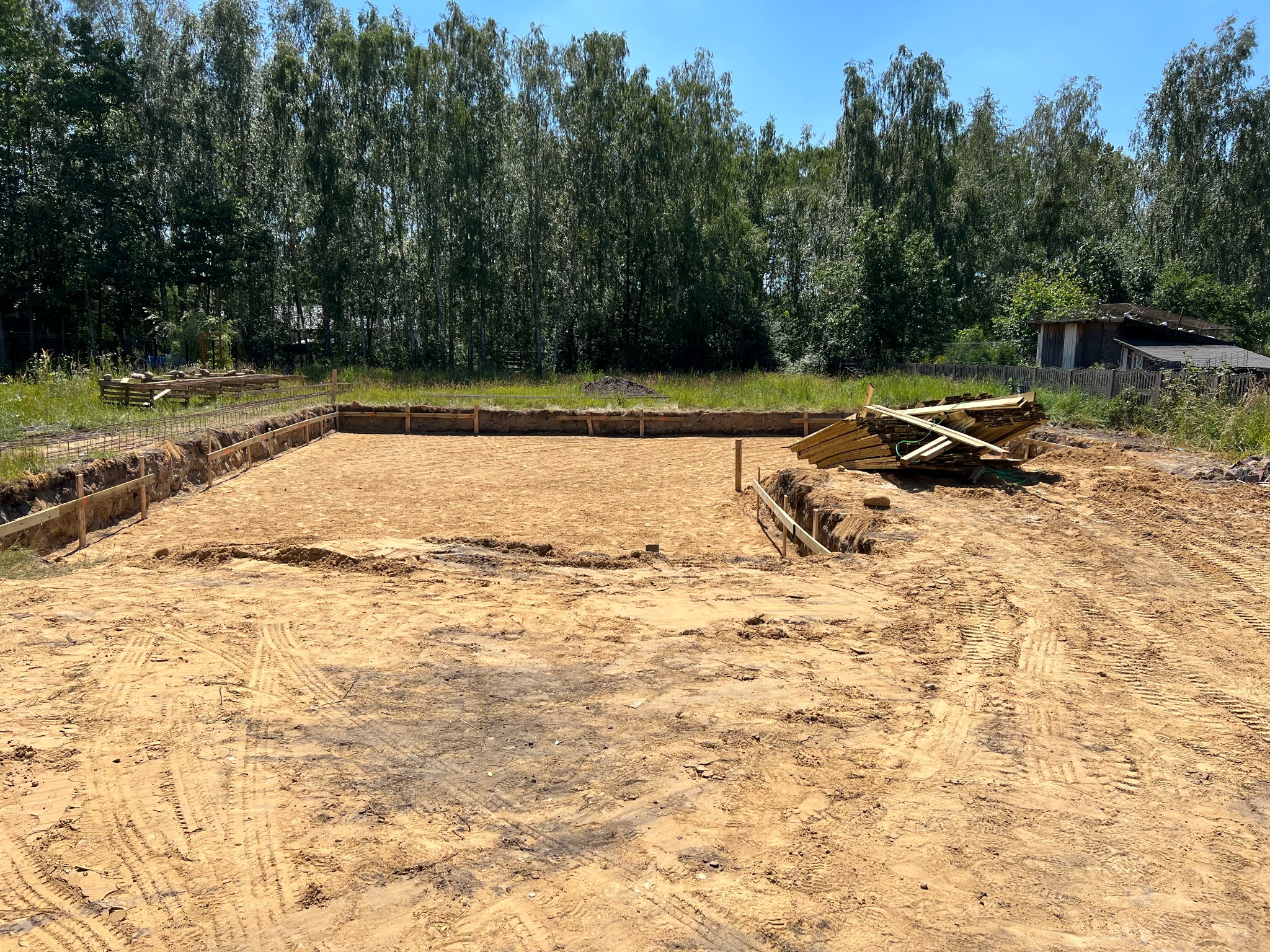 Przygotowany teren pod fundamenty domu jednorodzinnego w Grodzisku Mazowieckim z widocznymi wykopami, szalunkami i stertą desek. W tle drzewa i budynek gospodarczy.