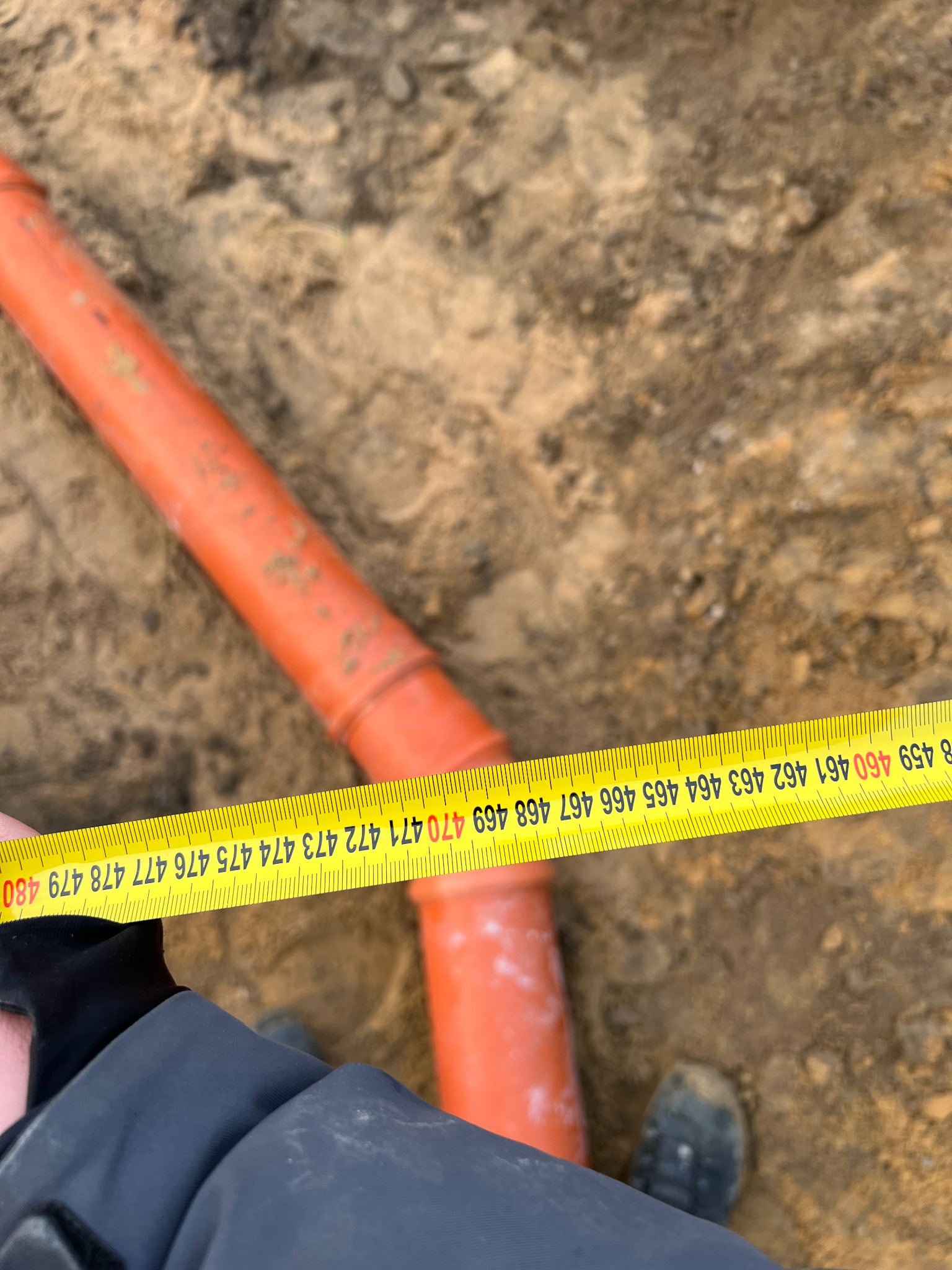 Pomarańczowa rura kanalizacyjna w wykopie mierzona żółtą taśmą mierniczą, widoczny fragment ubrania osoby mierzącej i but w ziemi.