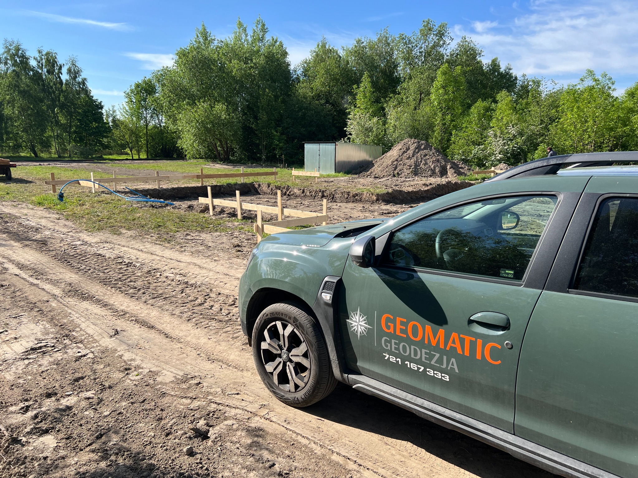 Geodezyjne wytyczenie fundamentów pod budowę domu, widoczny samochód terenowy z logo firmy Geomatic Geodezja, w tle drzewa i niebieski wąż do pomiarów.