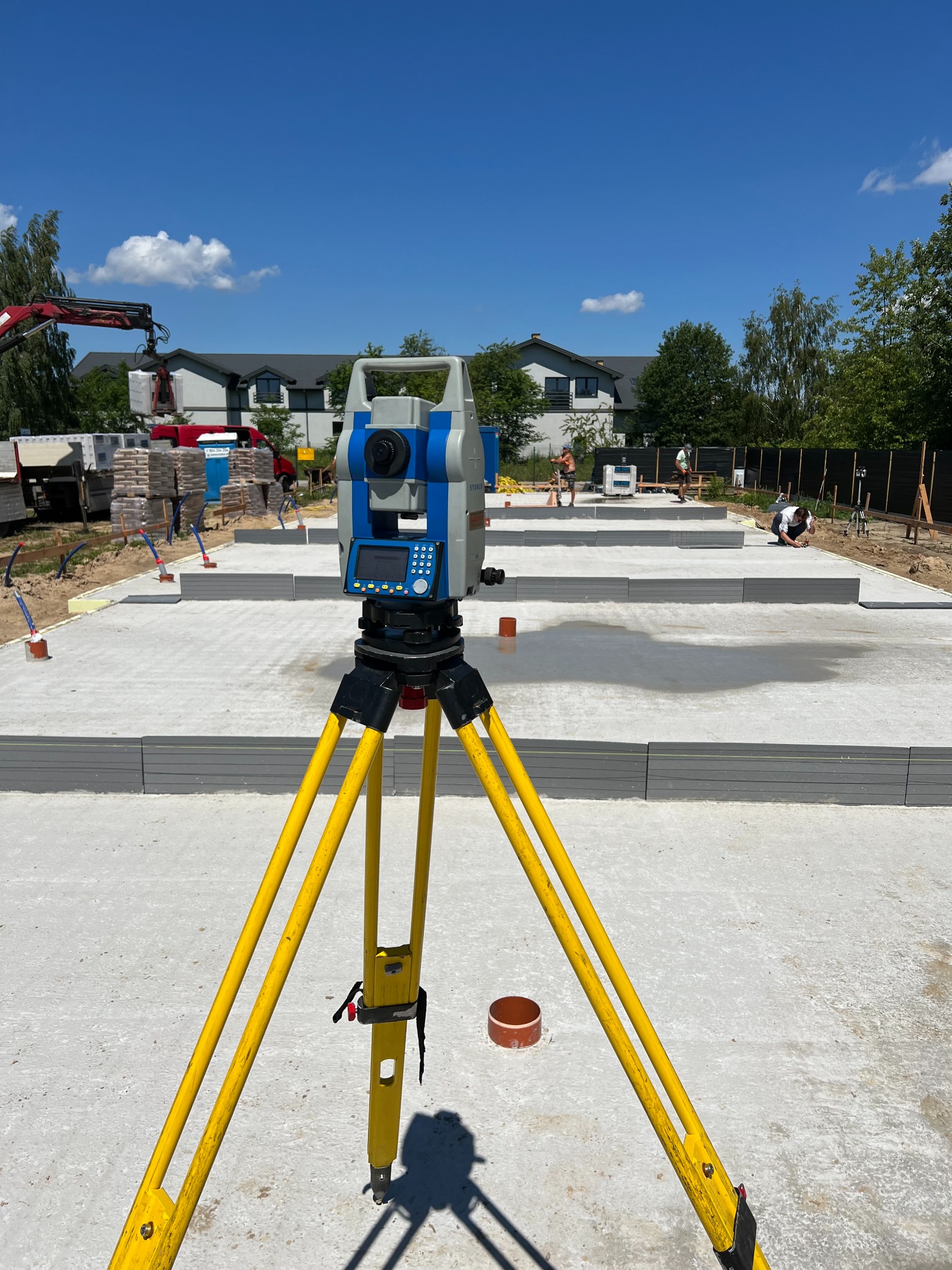 Żółty niwelator na trójnogu ustawiony na betonowej płycie fundamentowej w trakcie budowy domu jednorodzinnego, w tle pracujący robotnicy i dźwig podnoszący palety z cegłami.