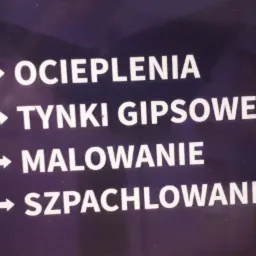 Tablica informacyjna z białymi napisami na ciemnym tle, wskazująca na usługi: Ocieplenia, Tynki Gipsowe, Malowanie, Szpachlowanie.