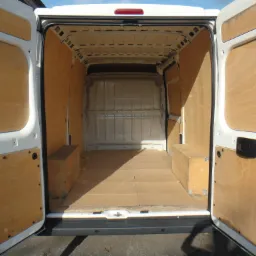 Wnętrze białego busa z otwartymi drzwiami, wyłożone sklejką i zabezpieczone kartonem na podłodze, z widocznymi drewnianymi skrzyniami po bokach.