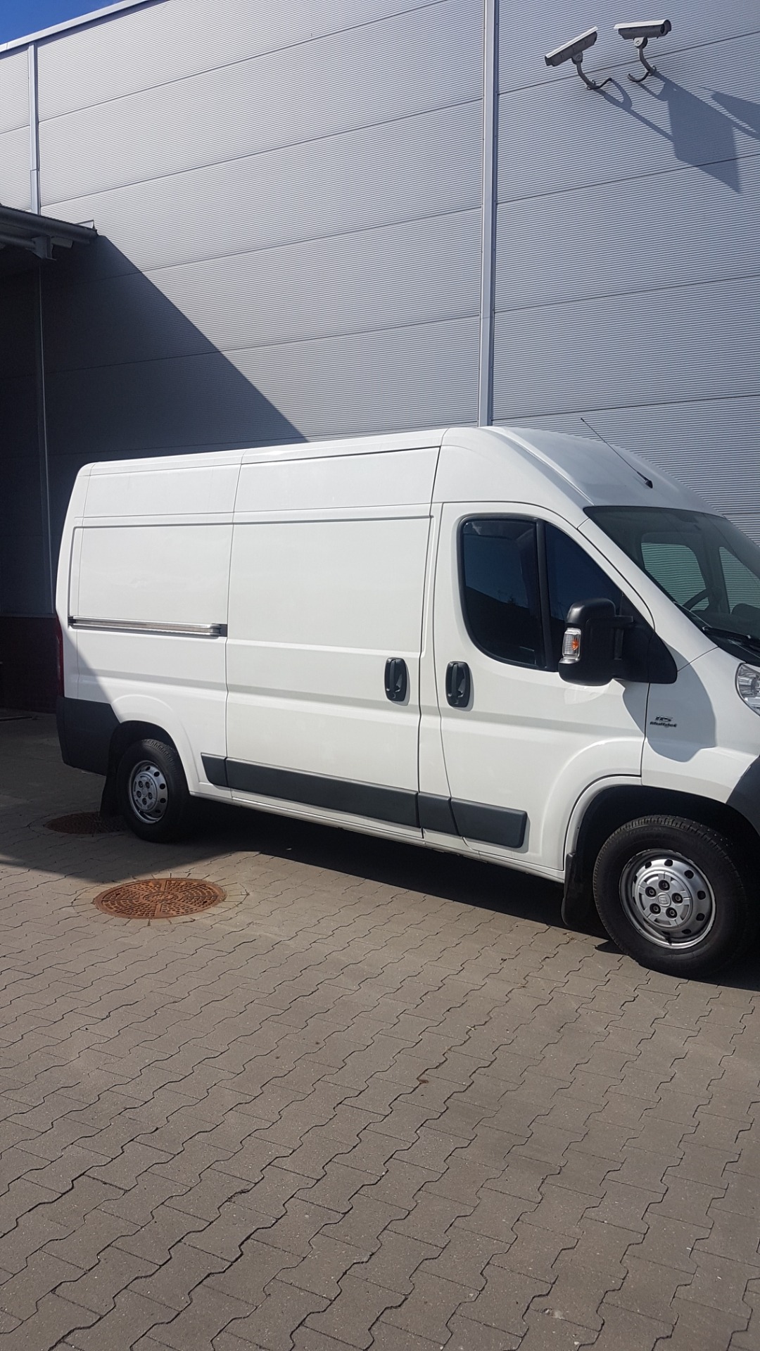 Biały bus dostawczy zaparkowany na kostce brukowej przed szarym budynkiem z monitoringiem.