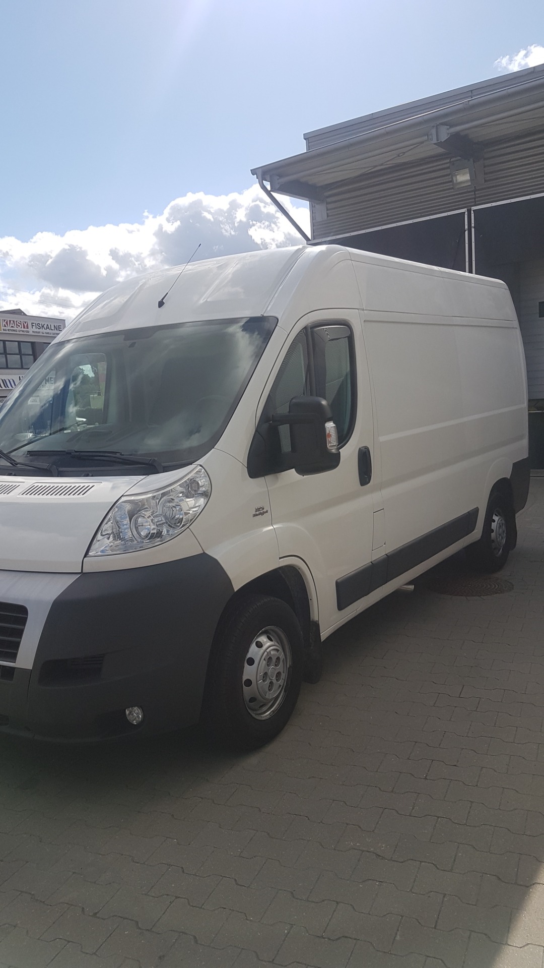 Biały samochód dostawczy marki Fiat Ducato na tle budynku z szarym dachem i błękitnego nieba z chmurami.