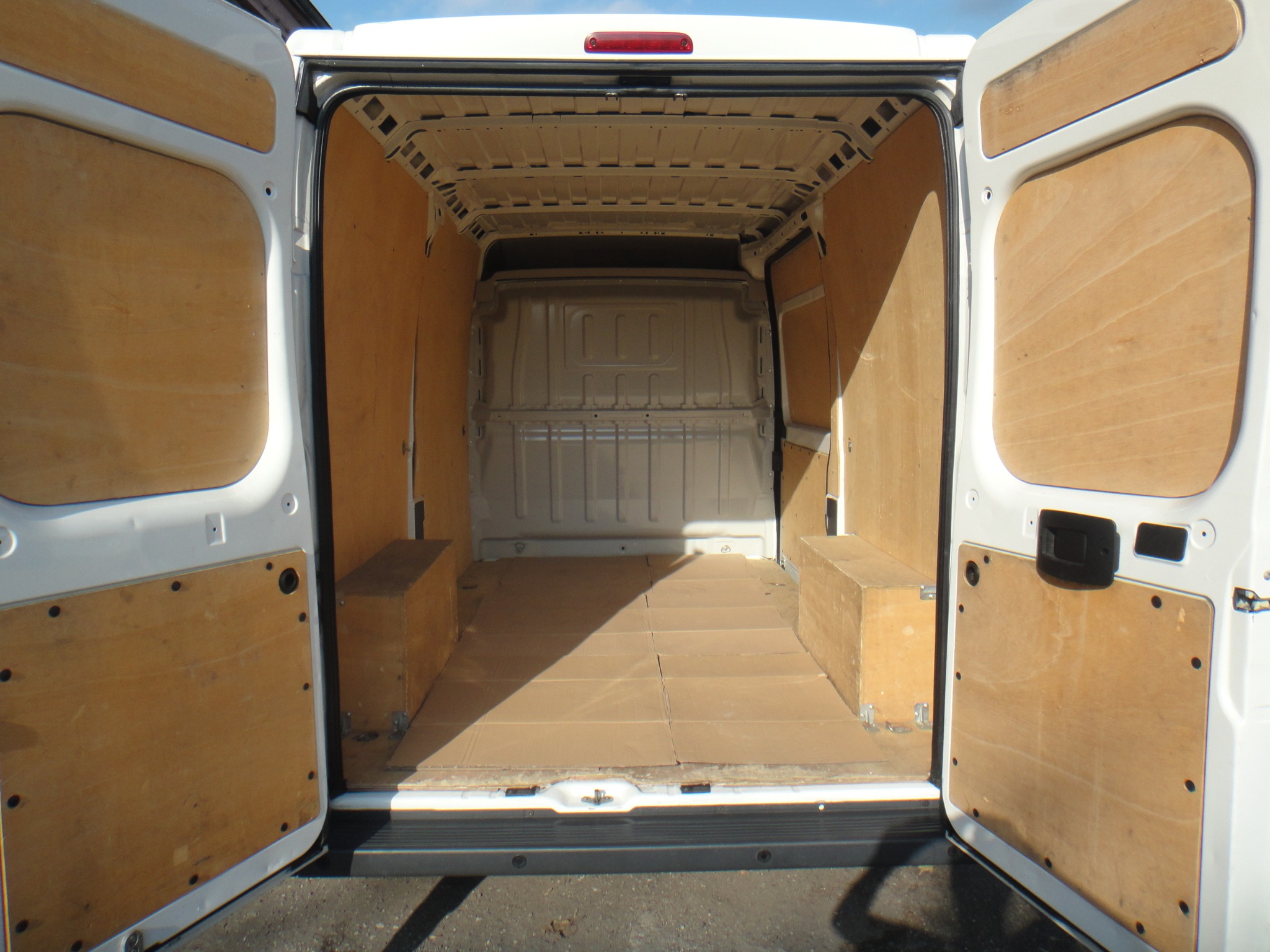 Wnętrze białego busa z otwartymi drzwiami, wyłożone sklejką i zabezpieczone kartonem na podłodze, z widocznymi drewnianymi skrzyniami po bokach.