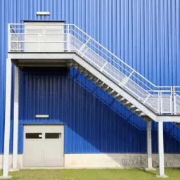 Zewnętrzne schody metalowe z podestem prowadzące do niebieskiej hali przemysłowej z blachy trapezowej, widoczne drzwi wejściowe i lampy oświetleniowe.