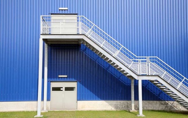 Zewnętrzne schody metalowe z podestem prowadzące do niebieskiej hali przemysłowej z blachy trapezowej, widoczne drzwi wejściowe i lampy oświetleniowe.