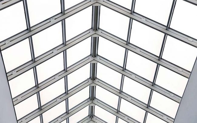 Szklany dach hali przemysłowej z widoczną stalową konstrukcją nośną, widok z dołu.