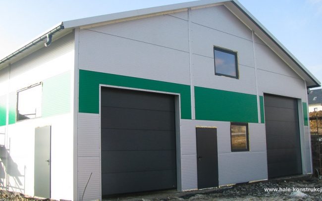 Zewnętrzny widok hali magazynowej z szarego panelu z dwoma czarnymi bramami segmentowymi i zielonym pasem dekoracyjnym, widoczne drzwi wejściowe i okna.