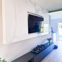 Lasak Projektowanie - Salon z minimalistyczną zabudową telewizyjną: biała marmurowa ściana, czarna szafka RTV z dekoracyjnym paleniskiem, drewniana podłoga i widok na ogród przez okno.