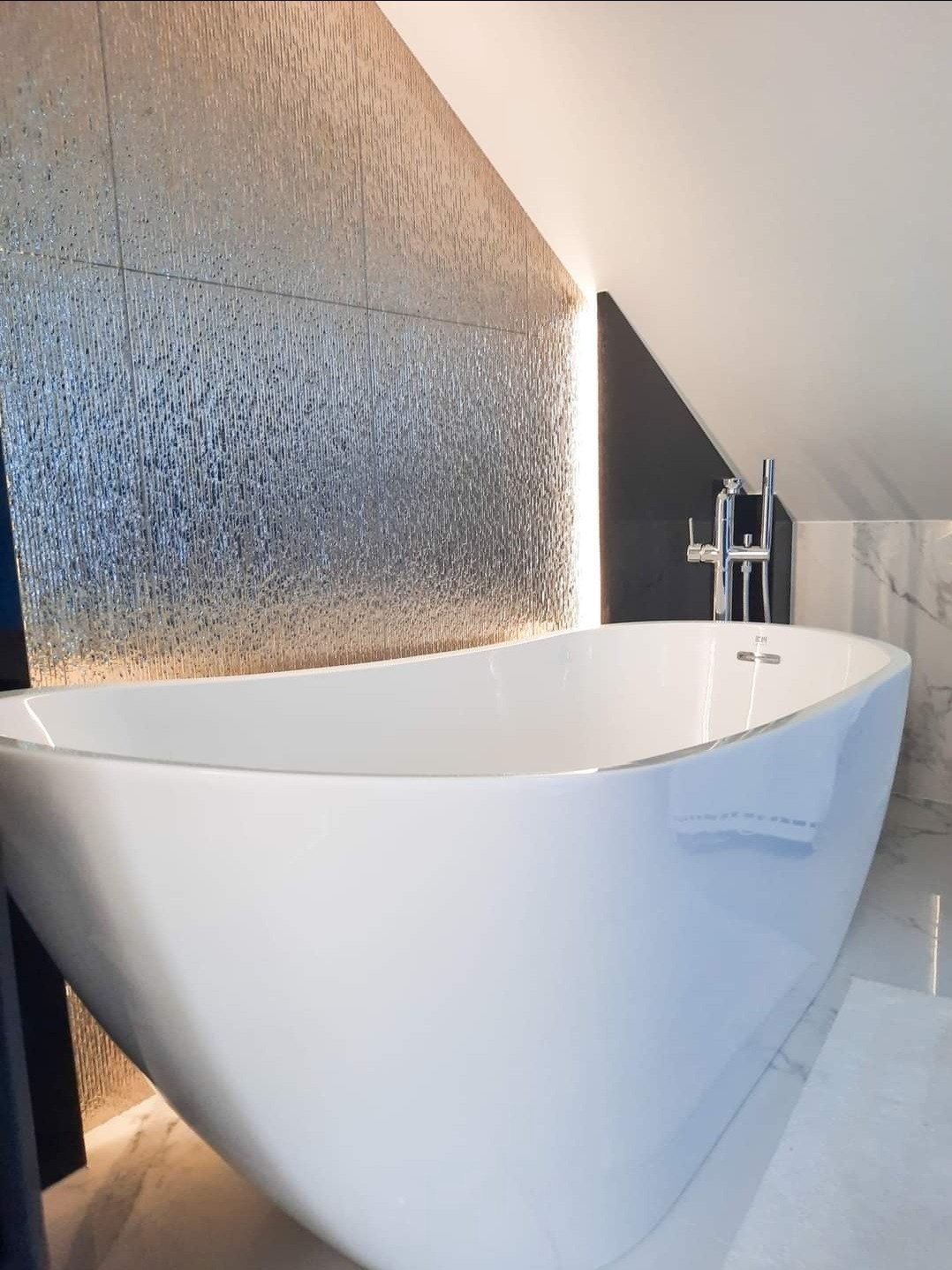 Wolnostojąca, biała wanna o owalnym kształcie pod skosem dachu, z chromowaną baterią, na tle ściany z dekoracyjnych, srebrnych płytek z subtelnym oświetleniem LED.