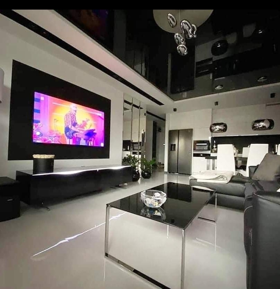 Nowoczesny salon z czarnym, połyskującym sufitem, dużym telewizorem w czarnej ramie, szklanym stolikiem kawowym i minimalistycznym oświetleniem.