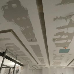 Finish line - Sufit podwieszany z płyt gipsowo-kartonowych w trakcie szpachlowania, widoczne profile i oświetlenie, drabina w tle.
