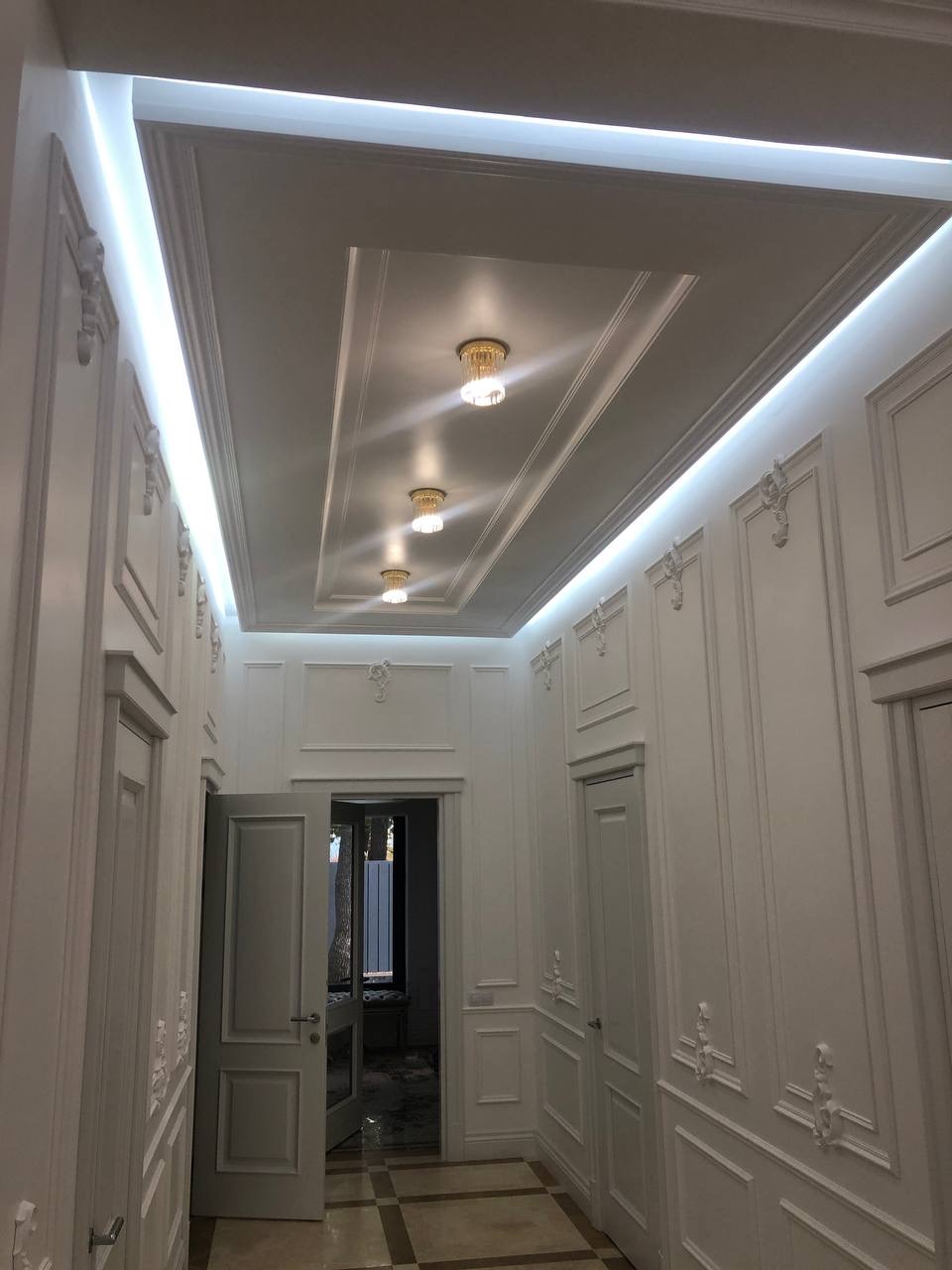 Korytarz w stylu klasycznym z białymi ścianami zdobionymi sztukaterią, podwieszanym sufitem z oświetleniem LED i trzema małymi, okrągłymi lampami, otwarte drzwi na końcu korytarza.