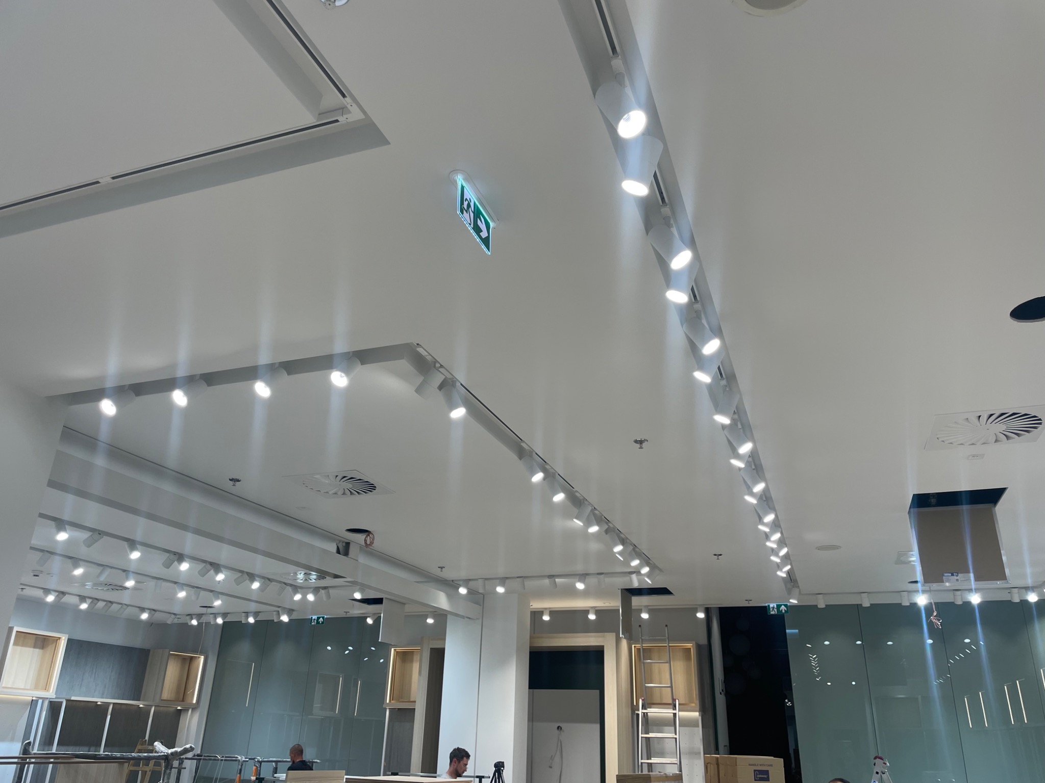 Montaż oświetlenia w biurze: widok na sufit z rzędami reflektorów punktowych, system wentylacji, znak ewakuacyjny i fragmenty zabudowy meblowej. W tle widoczni pracownicy.