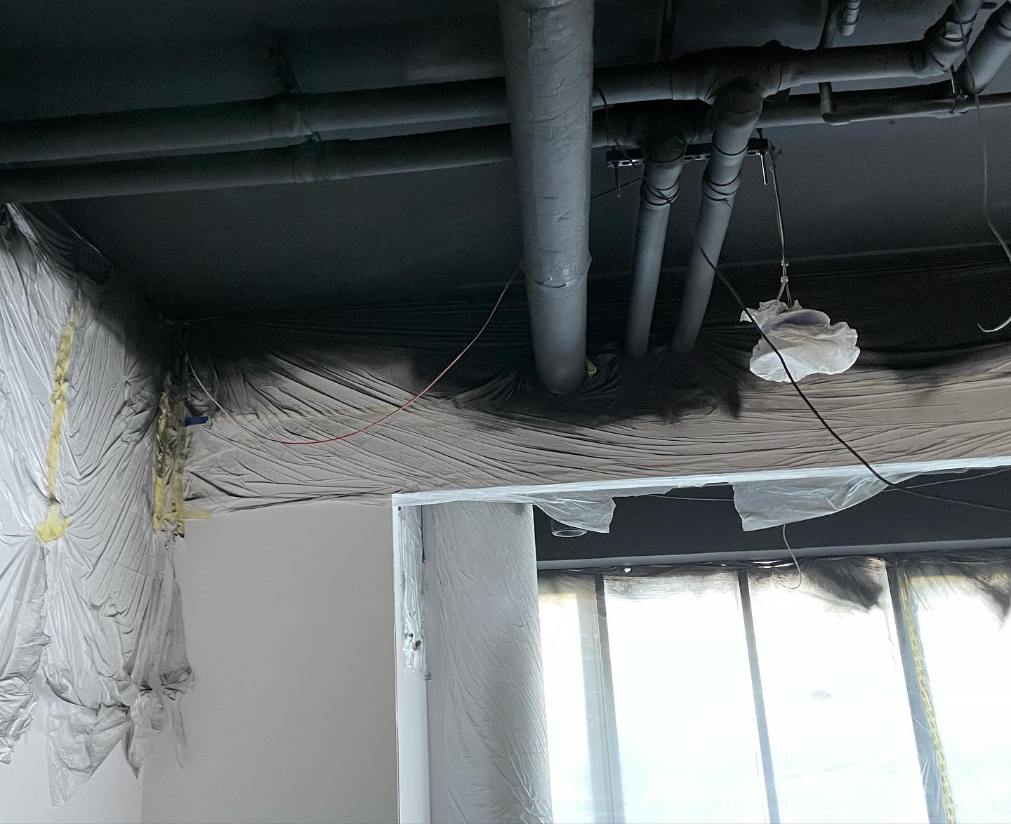 Przygotowanie pomieszczenia do malowania natryskowego: folia ochronna na ścianach, oknach i rurach pod sufitem, widoczne przewody elektryczne i fragmenty taśmy malarskiej.