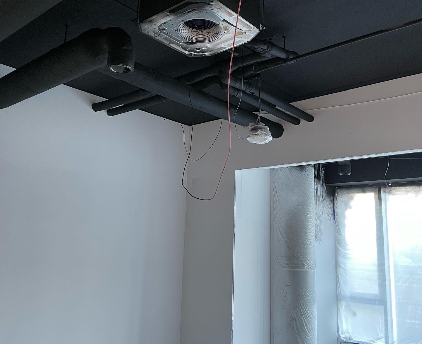Wnętrze w trakcie wykańczania: czarny sufit z widoczną instalacją wentylacyjną, białe ściany, okno zabezpieczone folią malarską, wiszące przewody elektryczne i zaklejona lampa.